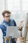 Чоловік дивиться на ноутбук під час малювання на мольберті — стокове фото