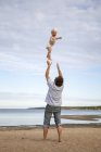 Homem jogando filho no ar na praia contra o céu com nuvens — Fotografia de Stock