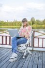 Женщина работает на ноутбуке на террасе возле реки — стоковое фото