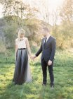 Наречений і наречений стоять на траві, тримаючись за руки, зосереджуючись на передньому плані — стокове фото