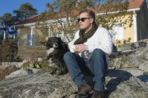 Мужчина сидит с собакой на скалах перед домом — стоковое фото