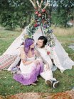 Bride and groom sitting on grass in front of white tent at hippie wedding — Stock Photo