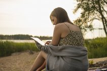 Young woman sitting on lakeshore and using digital tablet — Stock Photo
