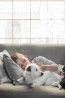 Menino deitado no sofá com cão perto da janela — Fotografia de Stock