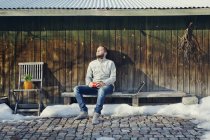 Середній дорослий чоловік сидить на лавці дерев'яним сараєм — стокове фото