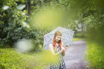 Жінка в квітковій сукні, використовуючи смартфон, стоячи з парасолькою в парку — стокове фото