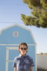 Хлопчик у сонцезахисних окулярах стоїть перед синім сараєм — стокове фото