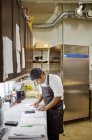 Man in apron writing in kitchen, differential focus — Stock Photo