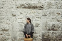 Young woman standing by stone wall holding basket — Stock Photo