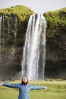Жінка стоїть з піднятими руками перед водоспадом — стокове фото