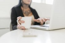 Tattooed woman holding mug while using laptop — Stock Photo