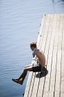 Молодой человек, сидящий на деревянной пристани у озера — стоковое фото
