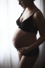 Беременная женщина смотрит вниз и стоит у окна — стоковое фото