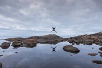 Rear view of woman jumping on rocks by lake under overcast sky — Stock Photo