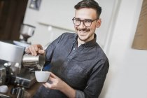 Barista sonriente haciendo café con leche y mirando a la cámara - foto de stock