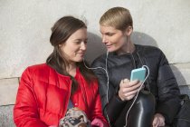 Дві жінки слухають музику на смартфоні, сидячи на сходах — стокове фото