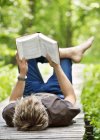 Молодий чоловік лежить на дерев'яній дрібці і читає книгу — стокове фото