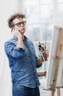 Man talking on phone next to easel and holding brushes — Stock Photo