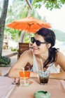 Mulher sorridente usando óculos de sol sentada com bebida no café — Fotografia de Stock
