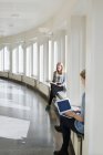 Young women reading book and using laptop by windows of university building — Stock Photo