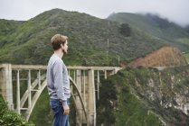 Man standing in front of bridge and mountains at Big Sur in California, USA — Stock Photo