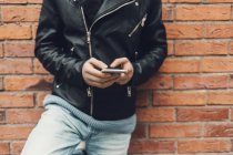 Cropped view of teenage boy leaning on brick wall, using smartphone, selective focus — Stock Photo