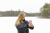 Giovane donna che prende selfie di fronte a un lago sulla neve — Foto stock