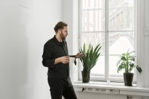 Mann mit Kopfhörer benutzt Smartphone und steht am Fenster — Stockfoto