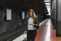 Junge Frau mit Koffer benutzt Smartphone an U-Bahnhof — Stockfoto