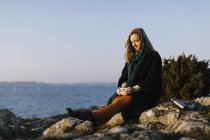 Donna che tiene la tazza seduta sulle rocce via mare — Foto stock