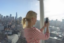 Femme adulte moyenne tenant un téléphone portable devant la fenêtre de l'appartement de la ville — Photo de stock