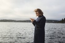 Jovem mulher de pé ao lado do lago e segurando telefone celular — Fotografia de Stock