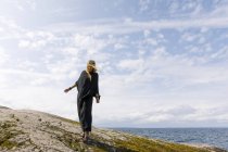 Femme balayée par le vent portant noir debout sur le rocher — Photo de stock