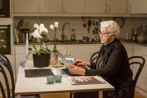 Старша жінка використовує ноутбук на кухні — стокове фото