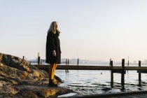 Femme debout sur les rochers par la mer pendant le coucher du soleil — Photo de stock