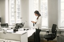 Mulher nova usando smartphone por mesa no escritório — Fotografia de Stock