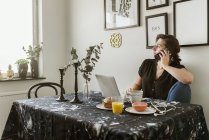 Молодая женщина использует ноутбук и смартфон во время завтрака — стоковое фото