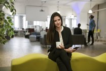 Junge Frau hält Smartphone mit Tasse in der Hand und lächelt in die Kamera, während sie im Büro auf dem Sofa sitzt — Stockfoto