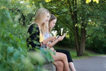 Сестры со смартфонами сидят на скамейке — стоковое фото