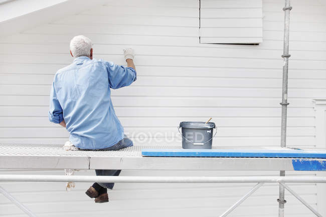 Вид ззаду літнього чоловіка, що сидить на риштуванні та малярській стіні — стокове фото