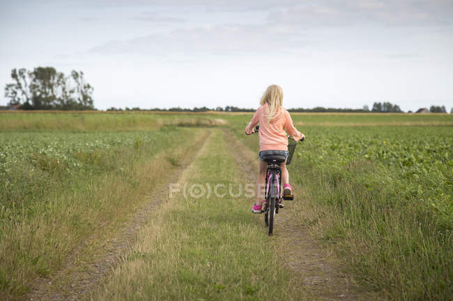 Blonde girl riding bike along dirt road in green field — Stock Photo