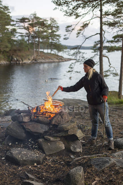 Mujer arrojando trozo de madera a la hoguera junto al lago al atardecer - foto de stock