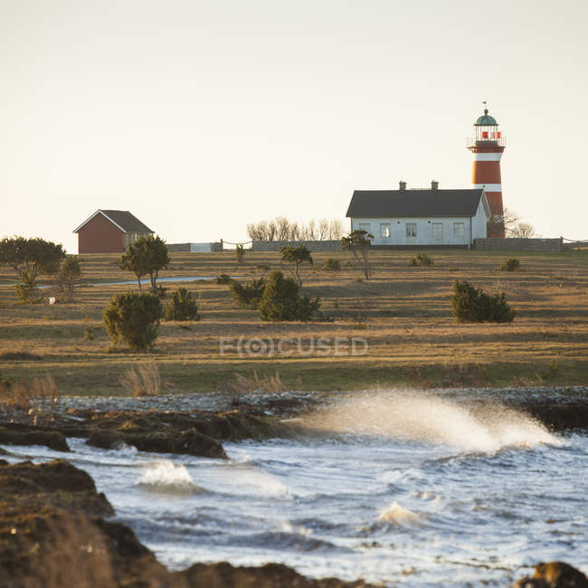 Surf waves on shore with small house and red lighthouse — Stock Photo