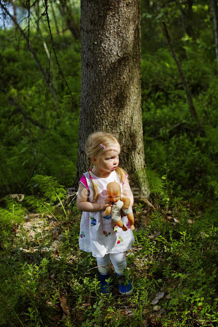 Girl with doll standing in front of trees — Stock Photo