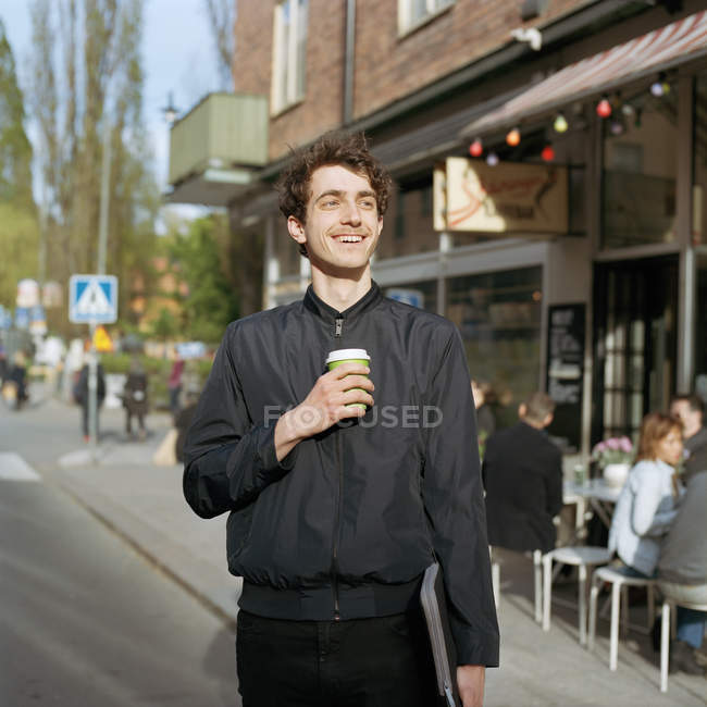 Улыбающийся мужчина, стоящий на улице с ноутбуком и одноразовой чашкой в руке — стоковое фото