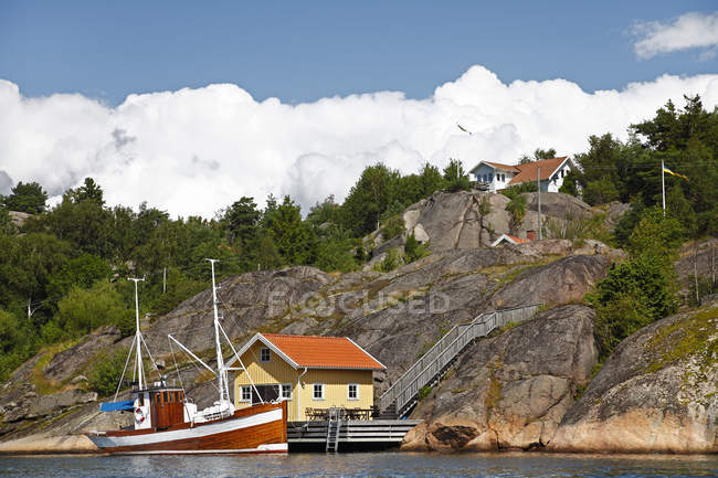 Barca ormeggiata vicino alla costa casa sotto cielo nuvoloso blu — Foto stock