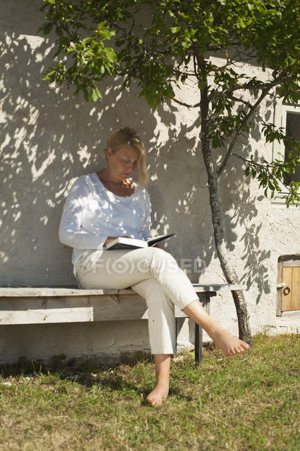 Woman sitting on backyard bench and reading book — Stock Photo