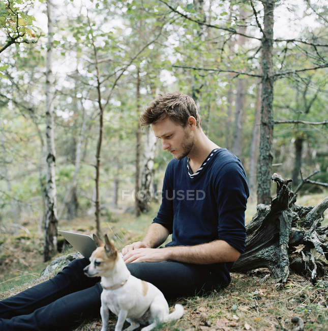 Чоловік використовує ноутбук біля собаки в лісі, вибірковий фокус — стокове фото