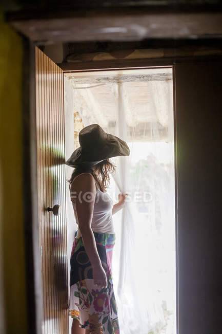 Femme en chapeau de soleil regardant à travers le rideau de porte — Photo de stock