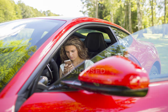 Дівчина-підліток сидить у червоній машині і використовує мобільний телефон — стокове фото